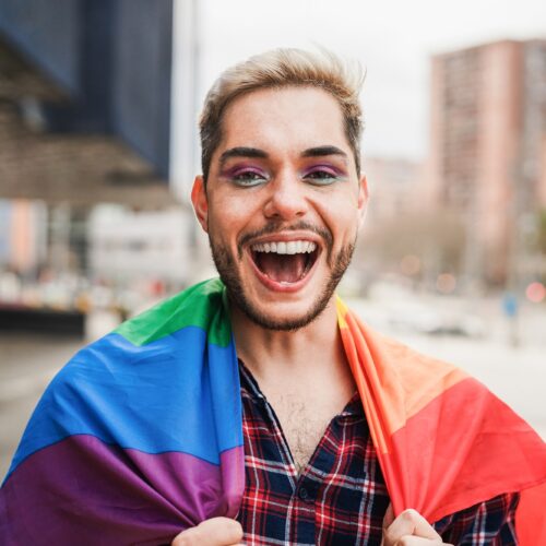 Gay man with makeup on having fun wearing lgbt rainbow flag outdoor - Focus on face
